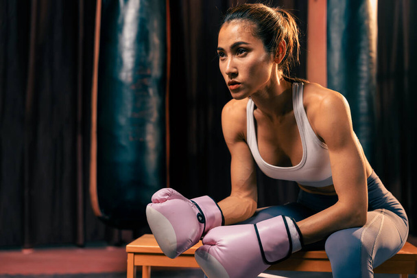 Ασιάτισσα γυναίκα Muay Thai πυγμάχος ή kickboxing λαμβάνοντας σύντομο διάλειμμα κάθεται με τα γάντια της στο γυμναστήριο με τον εξοπλισμό πυγμαχίας στο παρασκήνιο. Δυνατή και μυώδης αθλήτρια του σώματος. Ιμπέτους - Φωτογραφία, εικόνα