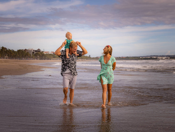 Feliz familia caucásica de padre, madre e hijo pasando tiempo en la playa. Padre llevando un niño en el cuello. Concepto de vacaciones de verano. Viajar a Asia. Playa de Seminyak, Bali, Indonesia - Foto, imagen