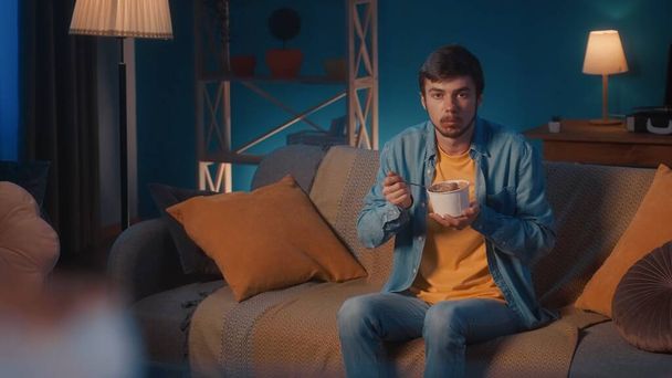 A man sits on a sofa in the living room and eats ice cream with a spoon from a bucket. A man with a full spoonful of ice cream looks straight ahead in surprise - Foto, immagini