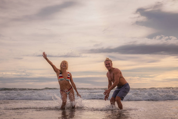 Glückliches kaukasisches Paar beim Wasserspielen am Strand. Lächelnd und lachend genießen junge Männer und Frauen das Leben. Sonnenuntergang. Bewölkter Himmel. Ozean mit Wellen. Urlaub in Asien. Bali, Seminyak, Indonesien - Foto, Bild