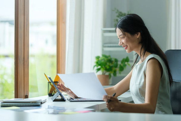 Schöne asiatische Geschäftsfrau benutzt Laptop-Computer und überprüft Finanzdokument am Schreibtisch. - Foto, Bild