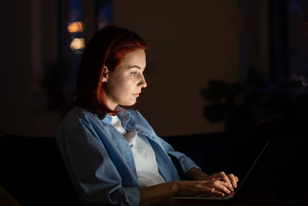 Gericht programmeur vrouw in donkere kamer zit op stoel werk op laptop. Workaholic freelancer Europese vrouw comfortabel zitten met computer op ronden werken aan grafisch ontwerp voor app 's nachts - Foto, afbeelding