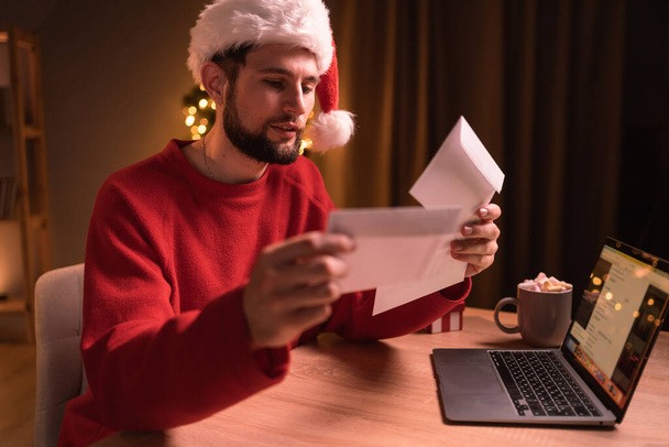 Worried men in Santa hat cheeking his mail late night in home office during christmas holidays. Copy space - Photo, Image