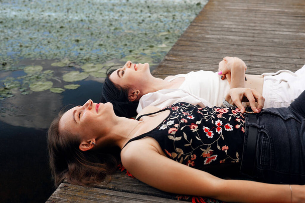 Két tinédzser lány ül egy mólón a folyóparton, és jól érzik magukat nyáron. boldog lány barátok pihentető szabadban tó közelében. - Fotó, kép