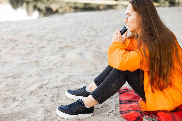 Молода жінка сидить на пляжі і курить електронну сигарету. Сумна дівчина в яскраво-помаранчевій куртці. - Фото, зображення
