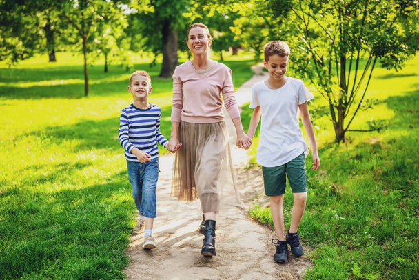 Happy mother is walking with her sons in park. They are holding hands and enjoying summer day. - Photo, Image