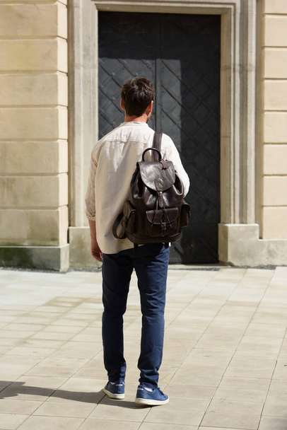 Handsome businessman walking on the street, with luxury leather backpack. Man wearing blue pants, sneakers, white t-shirt and beige shirt - Фото, изображение