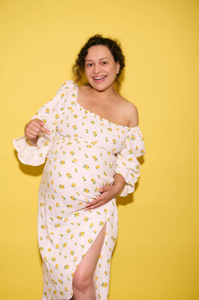 Verticaal studio portret van een vrolijke authentieke krullende multi-etnische gravitiste vrouw, aanstaande moeder wijzend naar haar zwangere buik, lachend vrolijk kijkend naar camera, geïsoleerde gele achtergrond - Foto, afbeelding