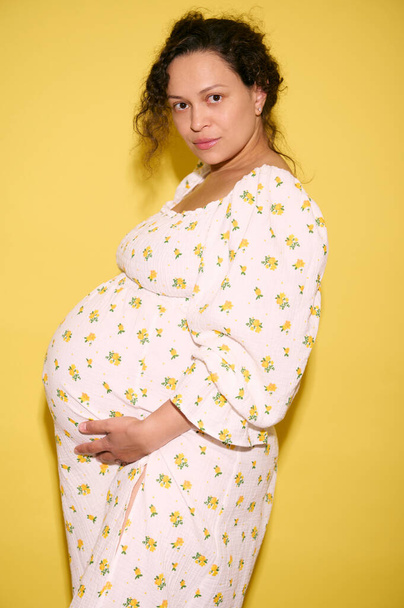 Αυτοπεποίθηση σοβαρή έγκυος σγουρά μαλλιά εθνοτική γυναίκα το καλοκαίρι sundress, αναμένοντας ένα μωρό, κρατώντας την κοιλιά της και κοιτάζοντας κάμερα. Άδεια μητρότητας. Εγκυμοσύνη Έννοια της τεκνοποίησης και της γέννησης - Φωτογραφία, εικόνα
