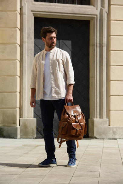 Όμορφος επιχειρηματίας περπατά στο δρόμο, με δερμάτινη τσάντα πολυτελείας. Άνδρας με μπλε παντελόνι, αθλητικά παπούτσια, λευκό t-shirt και μπεζ πουκάμισο - Φωτογραφία, εικόνα