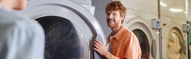 glimlachende jongeman in de buurt van wasmachines en vriendin in self service wasserij, banner - Foto, afbeelding
