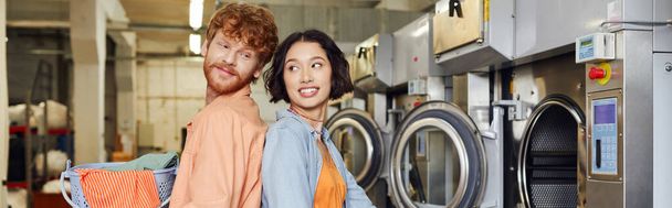 smiling man holding basket with clothes near asian girlfriend in coin laundry, banner - Foto, Bild