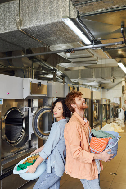 alegre pareja interracial sosteniendo cesta con ropa y de pie espalda con espalda en público lavandería - Foto, imagen