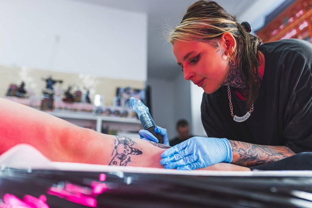 Skin art. Focused tattoo artist in protective gloves drawing a tattoo on a clients leg. Interior of a professional tattooing studio. High quality photo - Photo, Image