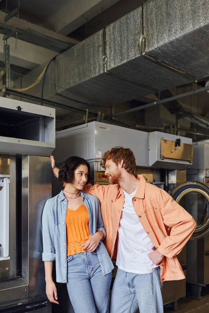 młody mężczyzna flirtuje z Azji dziewczyna w pobliżu pralki w publiczne pralnia - Zdjęcie, obraz