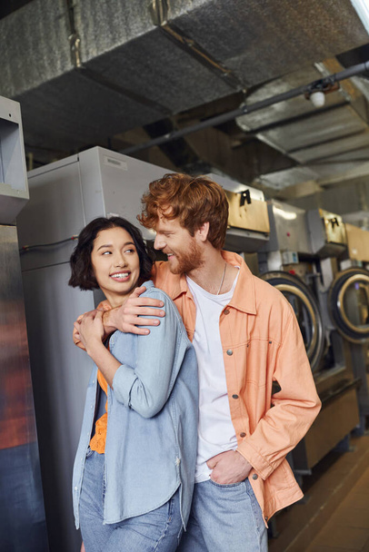 smiling young redhead man hugging asian girlfriend while standing in public laundry - Valokuva, kuva