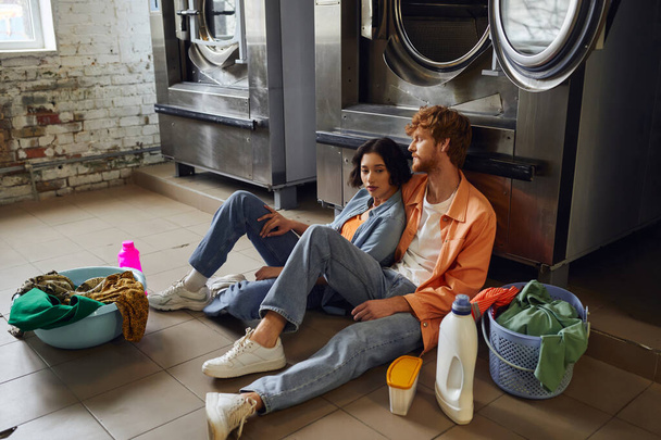 romantic multiethnic couple sitting near clothes and detergents on floor in public laundry - Photo, Image