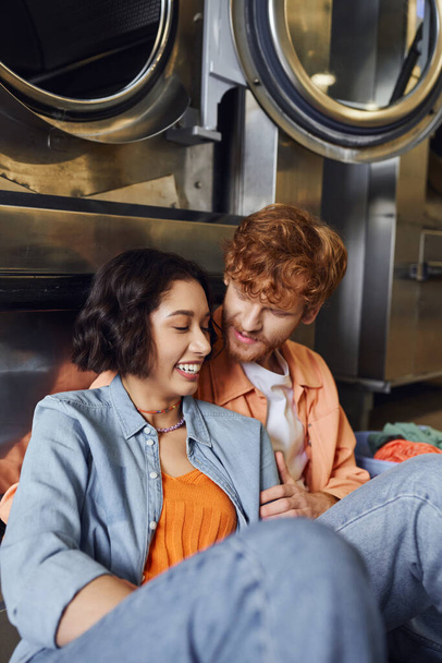 redhead man hugging cheerful young asian girlfriend near washing machine in coin laundry - Foto, Imagen