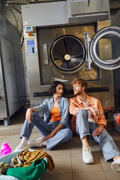 молодая межрасовая пара, сидящая рядом с одеждой и моющим средством на полу в общественной прачечной - Фото, изображение