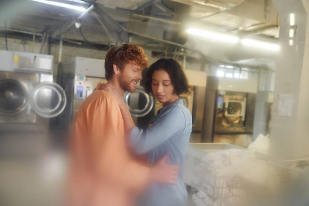 smiling young man hugging asian girlfriend while standing in blurred coin laundry - Photo, Image