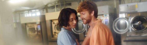 smiling young redhead man standing near brunette asian girlfriend in coin laundry, banner - 写真・画像