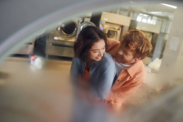 fiatal, többnemzetiségű pár ölelkezik, miközben elmosódott mosógép mellett állnak az érme mosodájában - Fotó, kép