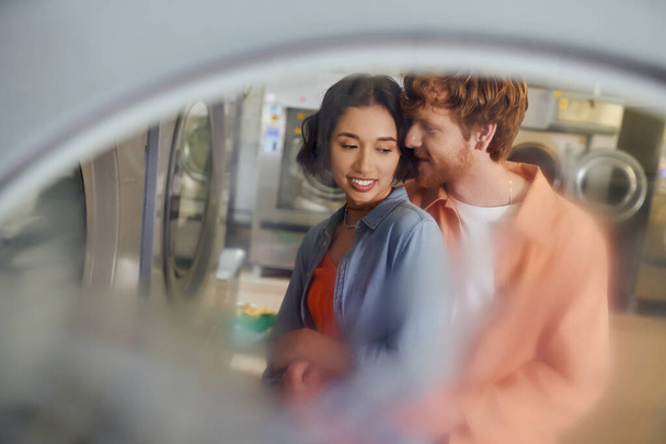 romantic redhead man hugging and flirting with asian girlfriend in public laundry - Фото, изображение
