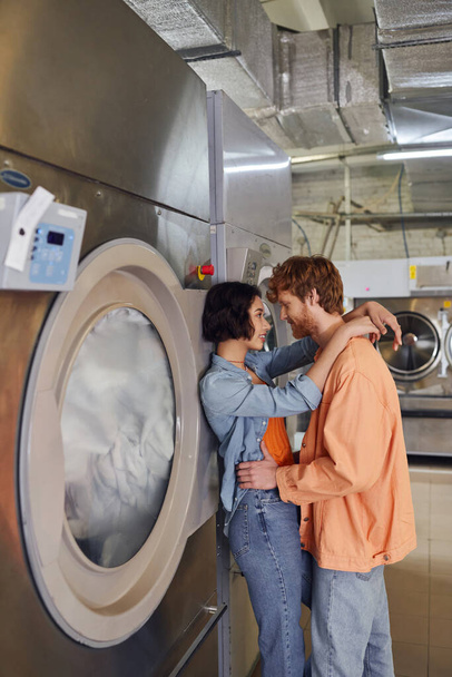 вид збоку усміхненої міжрасової пари обіймається, стоячи біля пральної машини в монетному пральні - Фото, зображення