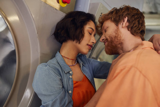 młoda wieloetniczna romantyczna para całuje się w pobliżu pralki w publicznej pralni - Zdjęcie, obraz