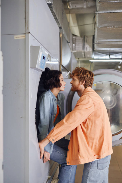 side view of smiling asian woman sitting on washing machine near romantic boyfriend in coin laundry - Photo, Image