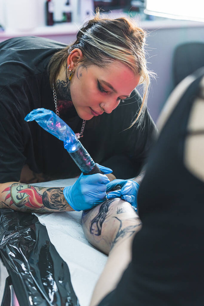 Вертикальний знімок художника жіночого татуювання під час її роботи. Робочий день у студії татуювання. Дівчина тримає татуювальну пістолет і малює татуювання на нозі. Високоякісна фотографія - Фото, зображення