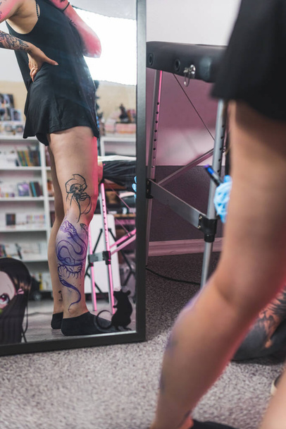 Vertical indoor shot of a caucasian person looking at their reflection of freshly drawn leg tattoo with freehand technique. High quality photo - Photo, Image
