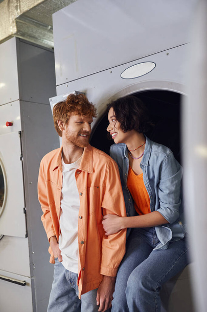 cheerful young asian woman hugging redhead boyfriend while sitting on washing machine in laundry - Zdjęcie, obraz
