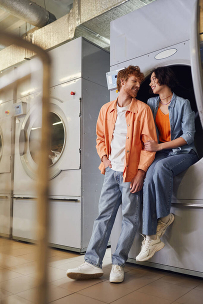 full length of cheerful multiethnic couple talking near washing machine in public laundry - Photo, Image