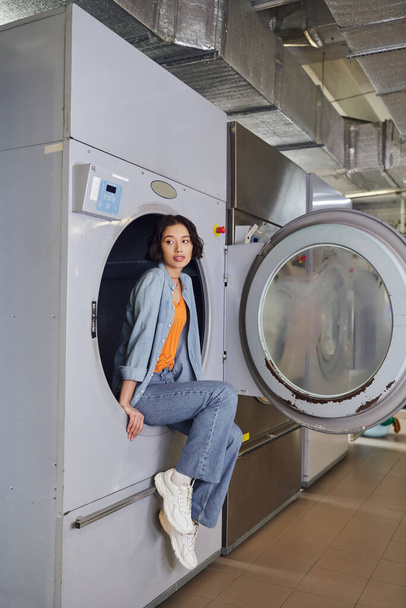 longitud completa de joven asiático mujer sentado en lavadora en público lavandería - Foto, Imagen
