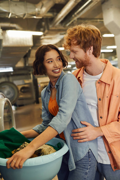 redhead man hugging cheerful asian girlfriend with clothes in basin in public laundry - Foto, immagini