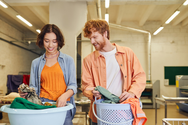 smiling young multiethnic couple putting clothes in baskets in public laundry on background - Photo, Image