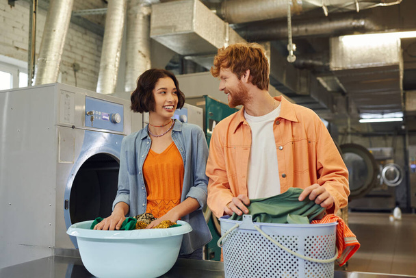 riendo interracial pareja hablando cerca de ropa en lavabos en público lavandería en fondo - Foto, imagen