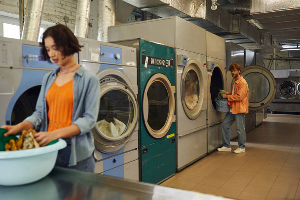 joven de pie cerca de la lavadora y novia borrosa con ropa en la lavandería de moneda - Foto, imagen