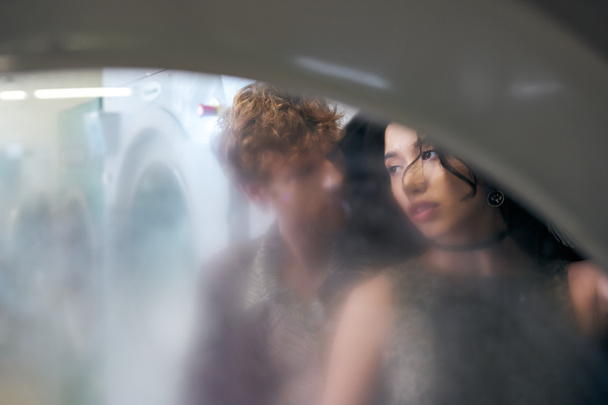 couple interracial dans des tenues élégantes debout derrière le verre de la machine à laver dans la blanchisserie à pièces - Photo, image