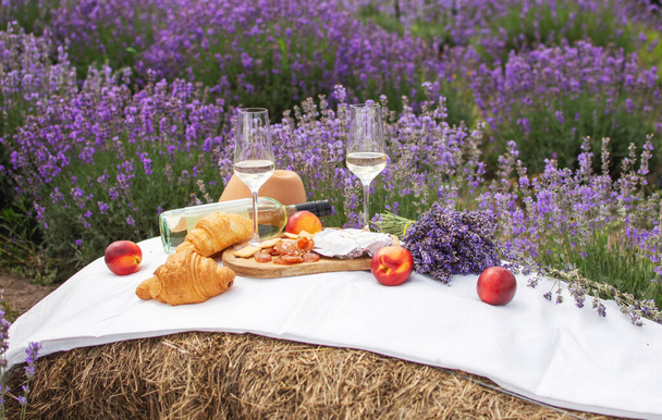Een zomerse picknick in een lavendelveld met croissants, perziken, salami, kaas en een fles wijn en glazen. Violet bloemen op de achtergrond. - Foto, afbeelding