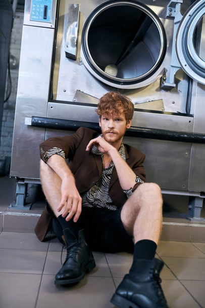 modischer Mann in Jacke und Shorts sitzt neben Waschmaschine in öffentlicher Wäscherei - Foto, Bild