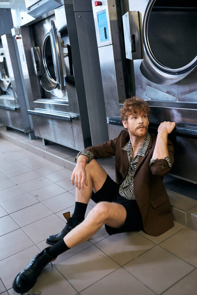 Çamaşırhanede çamaşır makinesinin yanında poz veren ceketli moda kızıl saçlı adam. - Fotoğraf, Görsel