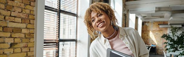 positive african american woman with braces smiling, holding folders in open space, startup, banner - Photo, Image
