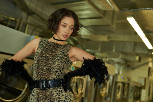 fashionable young asian woman in dress with animal print and gloves posing in coin laundry - Photo, Image