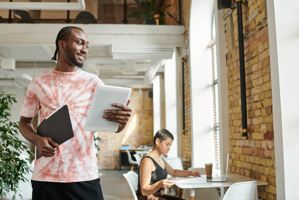 felice uomo africano americano in possesso di tablet, cartella, lavorando vicino a collega di lavoro femminile, progetto startup - Foto, immagini