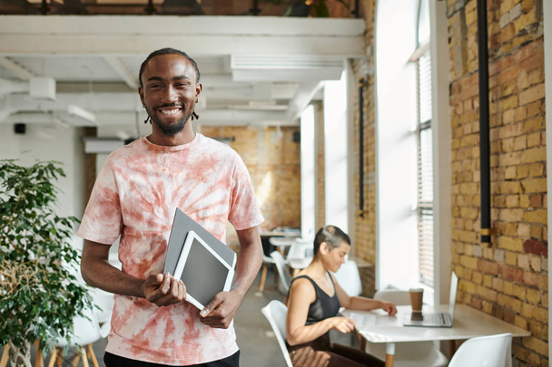hombre afroamericano feliz sosteniendo tableta, carpeta, mirando a la cámara cerca de compañera de trabajo, puesta en marcha - Foto, Imagen