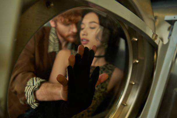 verschwommenes und stilvolles multiethnisches Paar, das Glas Waschmaschine in Münzwäsche berührt - Foto, Bild