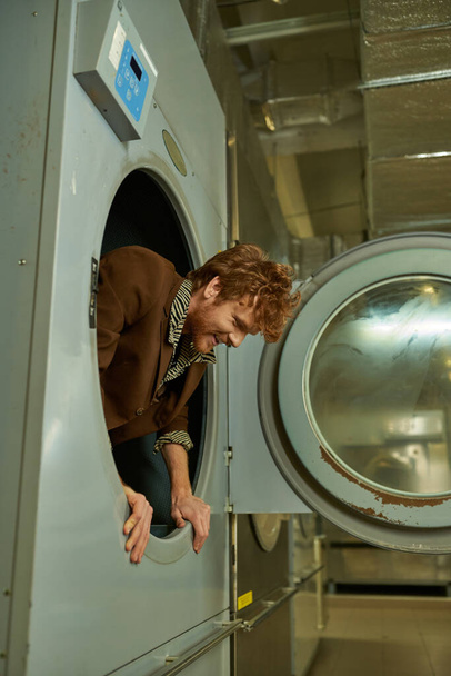 весёлый и модный молодой рыжий мужчина позирует в стиральной машине в публичной прачечной - Фото, изображение