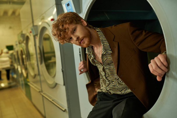 confident young redhead man looking at camera while posing in washing machine in coin laundry - Photo, Image